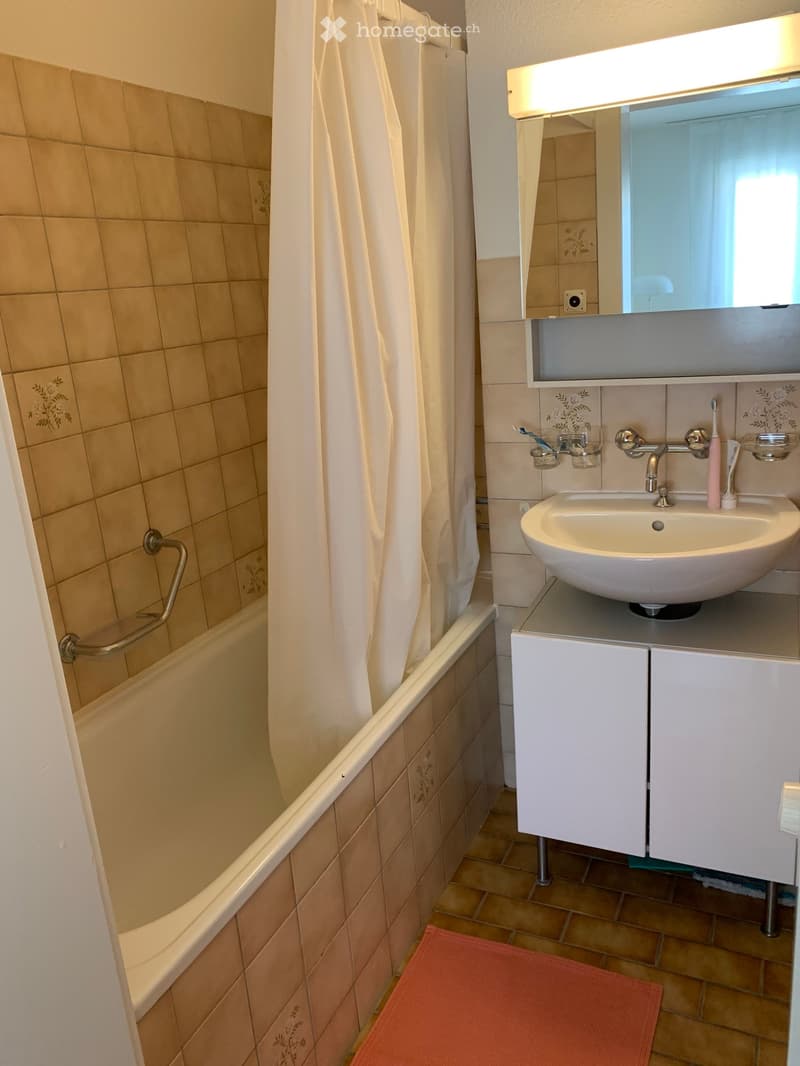 Bathroom with a bathtub, sink, mirror, and cabinet. Beige tiles and a red bathroom mat are visible.