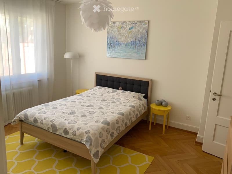 Cozy bedroom with wooden flooring, a bed with a leaf-patterned duvet, a nightstand with vases, a floor lamp, and a painting on the wall.