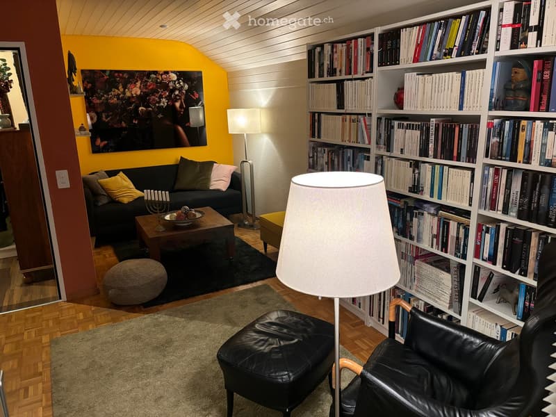 Living room with a black sofa, coffee table, bookshelf filled with books, two black leather chairs, a floor lamp, and a table lamp. The room features wooden flooring and a yellow accent wall.
