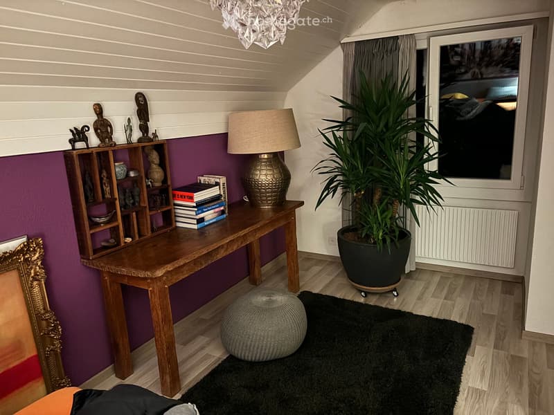 A modern, cozy living space with a wooden table, a large potted plant, a round stool, a shelf with decorative items and books, and a stylish lamp.