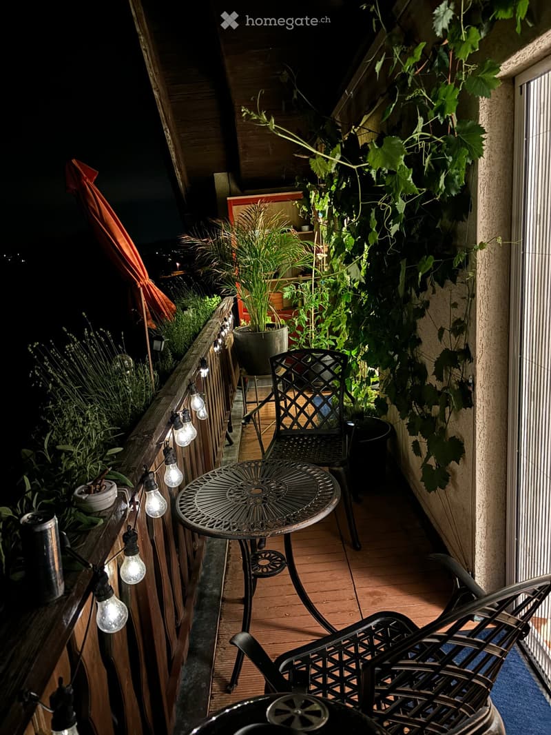 Balcony with wooden decking, string lights, metal table and chairs, potted plants, and a vine climbing on the wall.