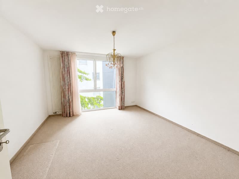 Empty room with white walls, carpeted floor, large window with curtains, and a chandelier hanging from the ceiling.