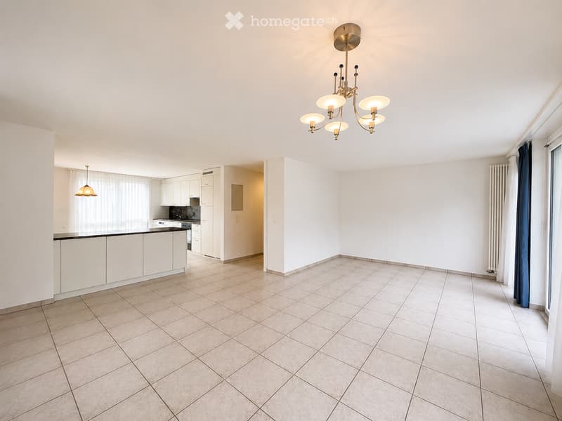 Bright, open living space with tiled flooring, white walls, and a modern kitchen area featuring white cabinets and a countertop. A chandelier hangs from the ceiling, and a radiator is visible along the wall.