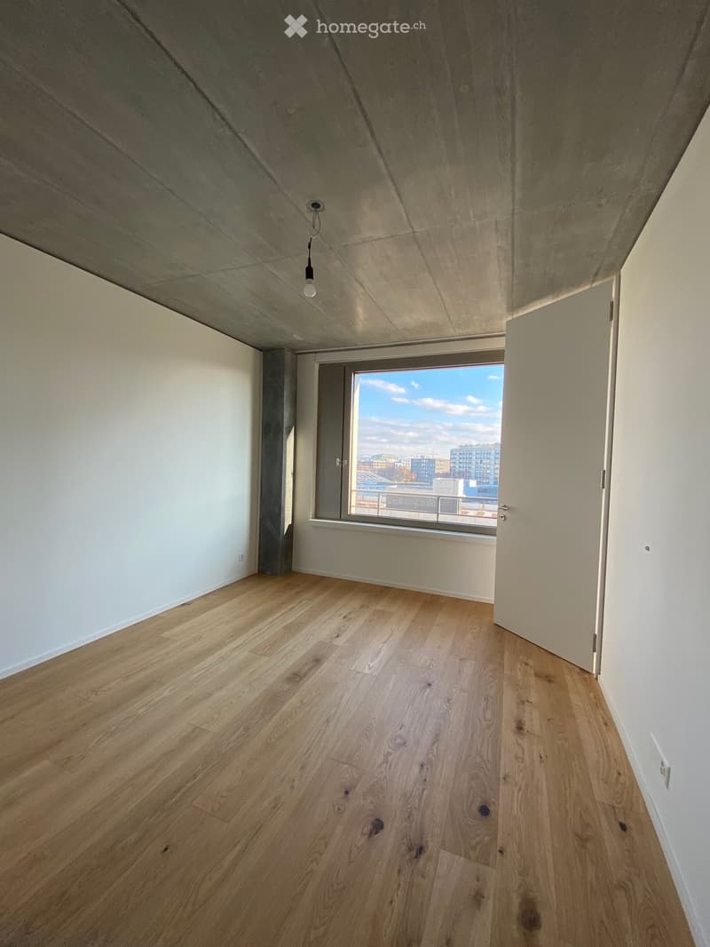 Empty room with light wooden flooring, white walls, and a large window offering a city view.