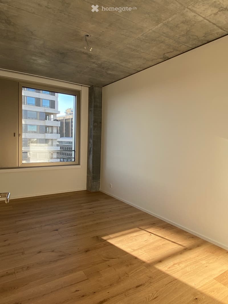 Empty room with wooden flooring, white walls, and a large window showing a view of a building outside.