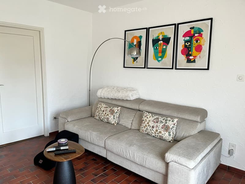 Living room with a gray sofa, a wooden table, a floor lamp, three paintings on the wall, and a white door.