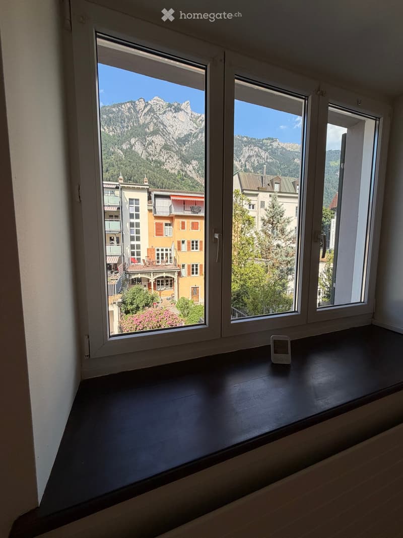 Empty room with large windows offering a scenic mountain view and townscape. A small device is placed on the windowsill.