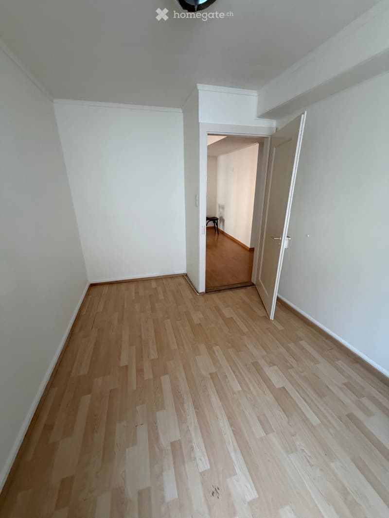Empty hallway with light wooden flooring and white walls, featuring an open door leading to another room.