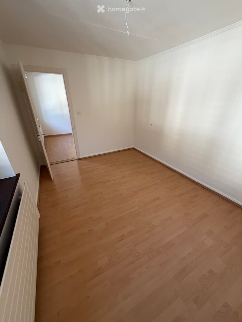 Empty room with light wooden flooring and white walls, featuring a partially open door leading to another room.