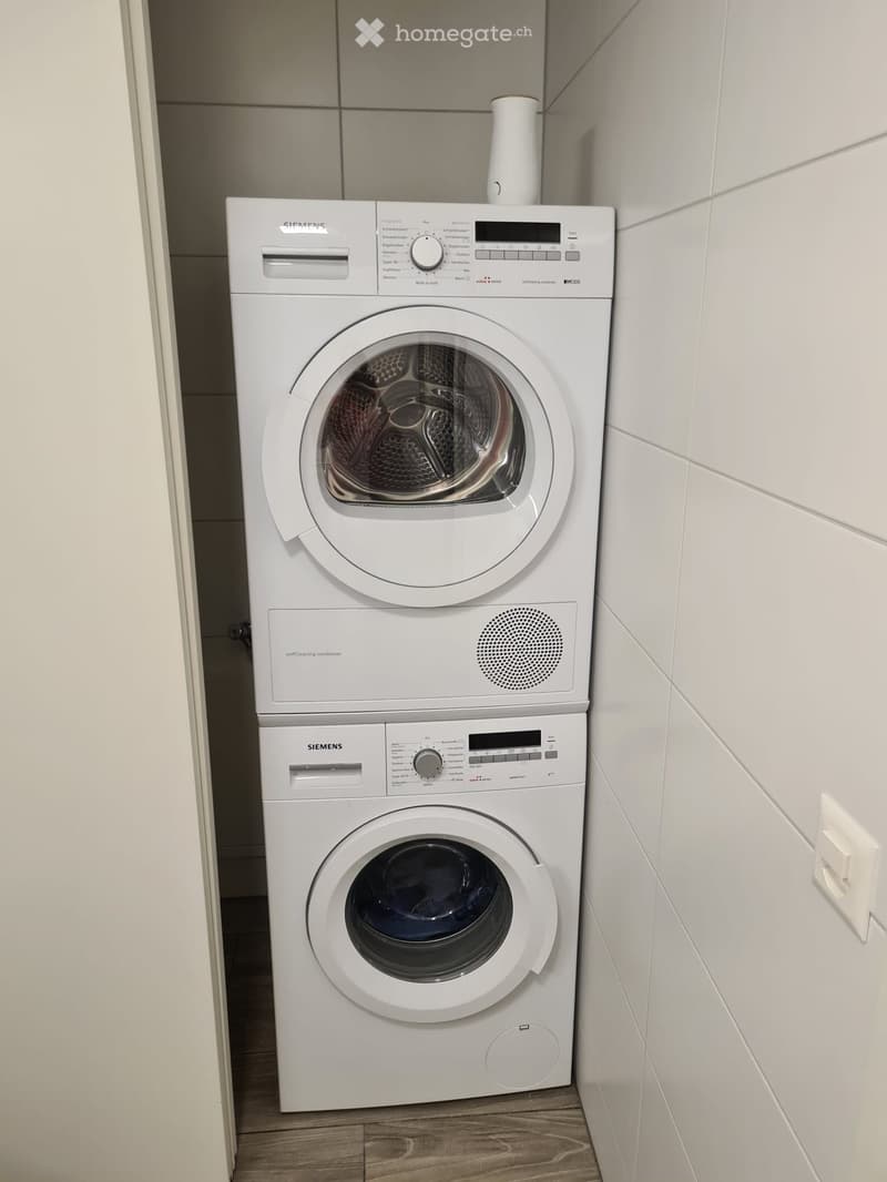 White Siemens washing machine and dryer stacked vertically in a tiled bathroom with wooden flooring.