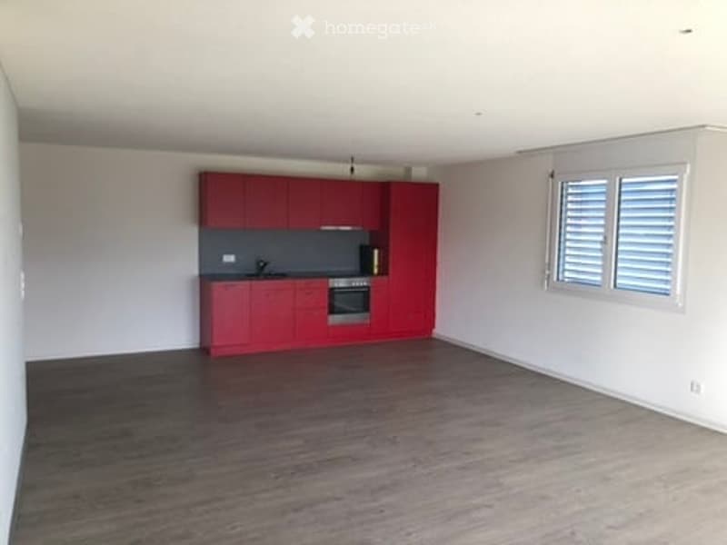 Empty room with red kitchen cabinets, a sink, and a window with shutters. The floor is wooden, and the walls are white.