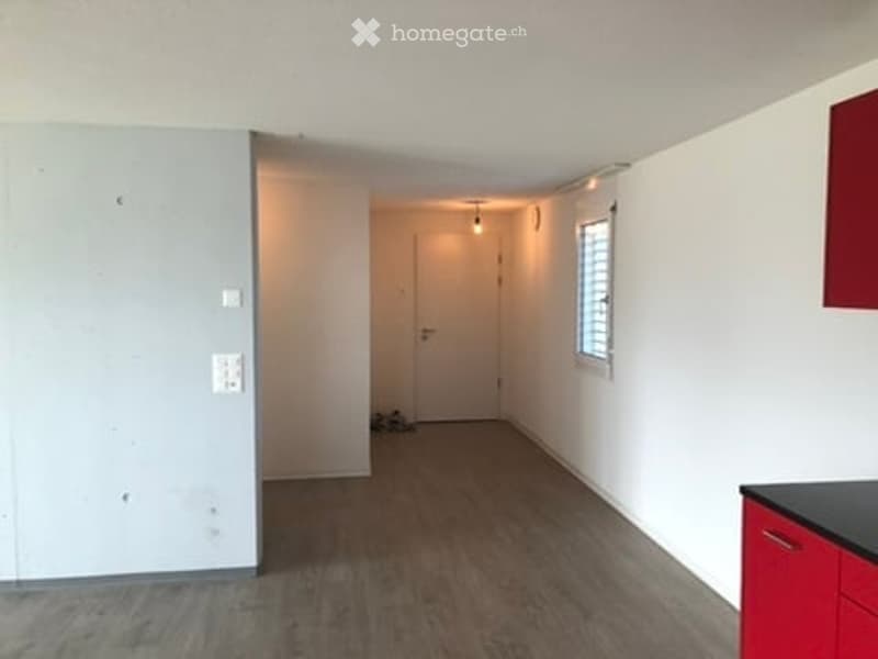 Empty room with white walls, wooden flooring, and a small window. There is a red cabinet visible on the right side.