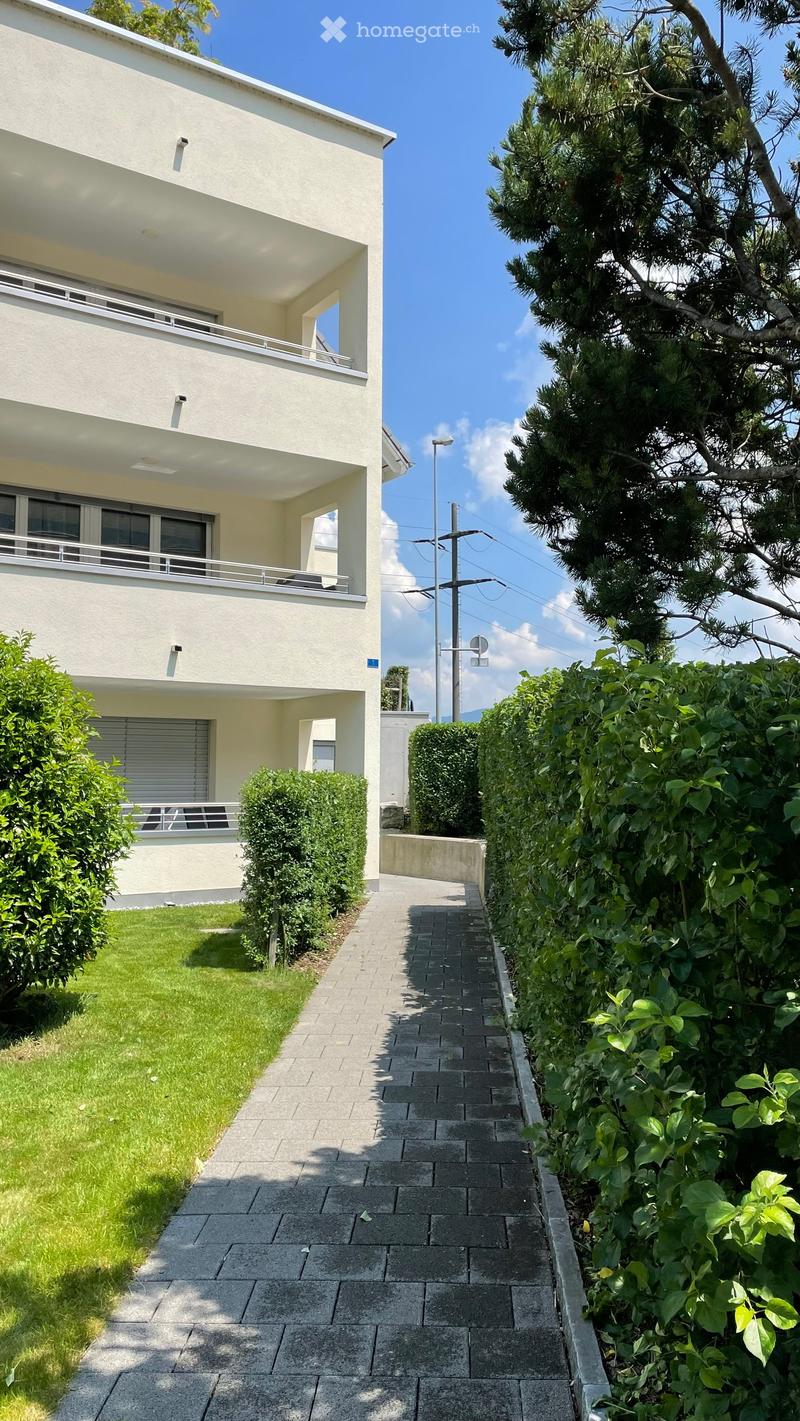 Modern two-story building with white facade, multiple windows, and balconies, surrounded by neatly trimmed hedges and a paved pathway.