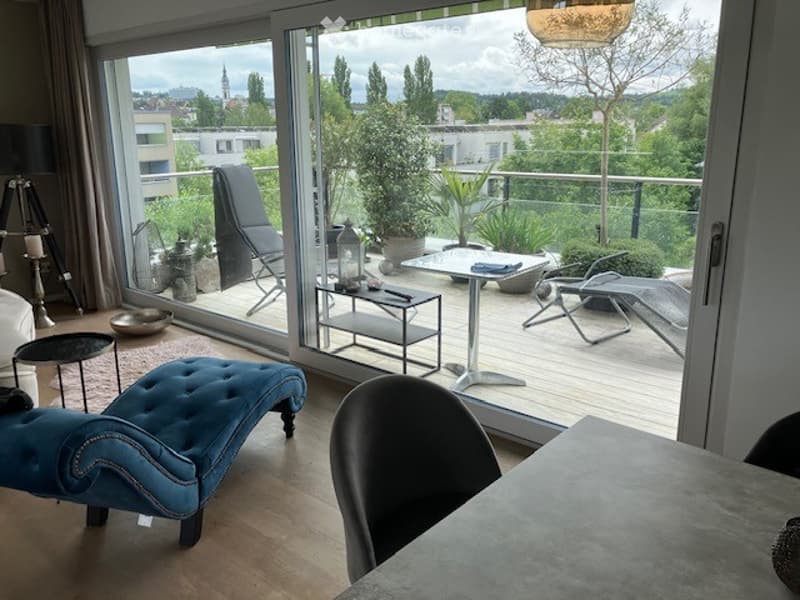 A modern living room with a large glass balcony door leading to a wooden deck. The room features a blue tufted sofa, a black dining chair, a small table, and a floor lamp. The balcony has outdoor furniture including a lounge chair, a small table, and pott
