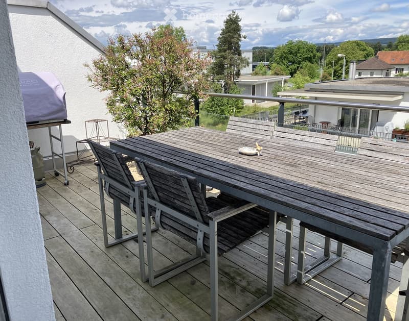 Balcony with wooden table, chairs, gas cylinder, and grill, overlooking a green landscape.