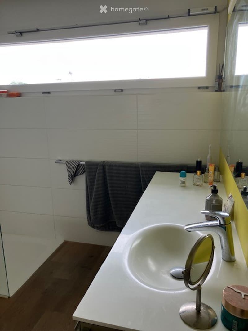 Modern bathroom with a white sink, chrome faucet, mirror, and various toiletries on the counter. A dark towel hangs on a rack, and the walls are tiled in white.