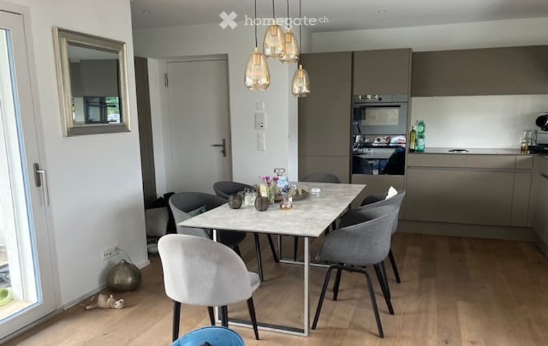 Modern, open-plan kitchen and dining area with light wood flooring, grey chairs, a white dining table, pendant lights, and kitchen cabinets.