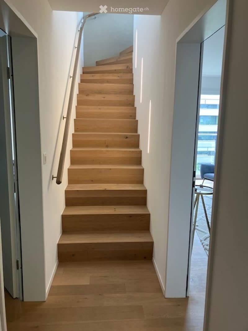 Modern wooden staircase with a sleek metal railing, leading up to an upper level. The hallway features light-colored wooden flooring and white walls.