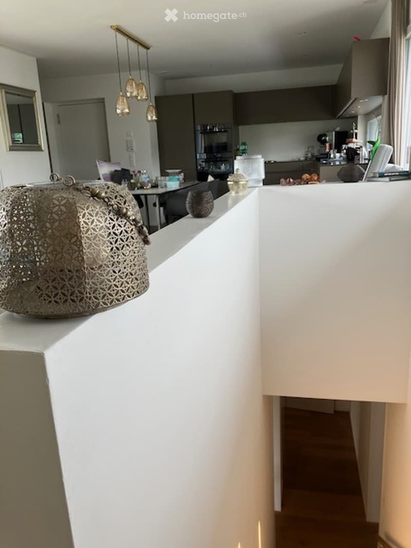 Modern kitchen with white countertops, dark cabinets, and pendant lights. A handbag is placed on the counter, and a small table with chairs is visible in the background.