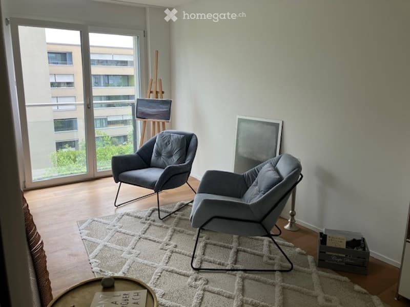 Modern living room with large windows offering a city view, featuring a grey sofa, two armchairs, a coffee table, and an art piece on the wall.