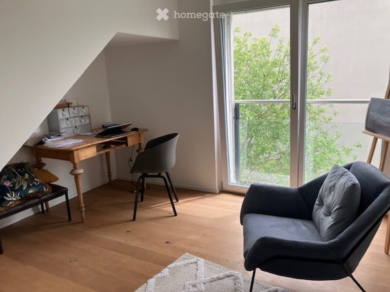 Bright, modern room with wooden flooring, large windows, and a balcony. Features a desk with a laptop, a comfortable sofa, and a cozy chair.