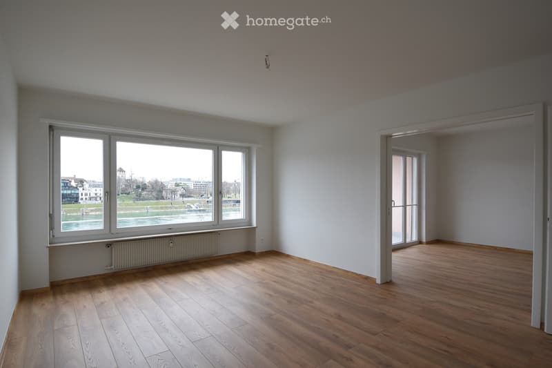 Empty room with wooden flooring, large windows overlooking a river, and a radiator beneath the windows.