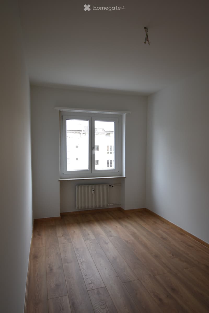Empty room with wooden flooring, white walls, a window with white frame, and a radiator below the window.