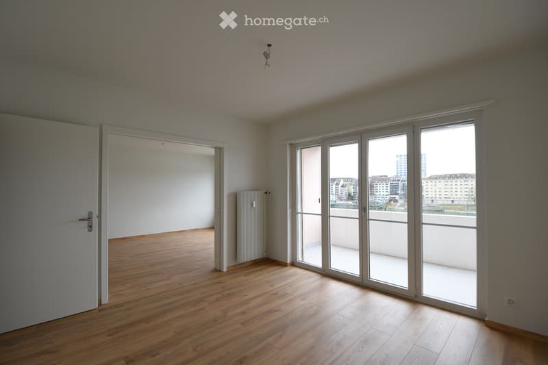 Empty room with wooden flooring, white walls, large sliding glass doors leading to a balcony, and a radiator on the wall.