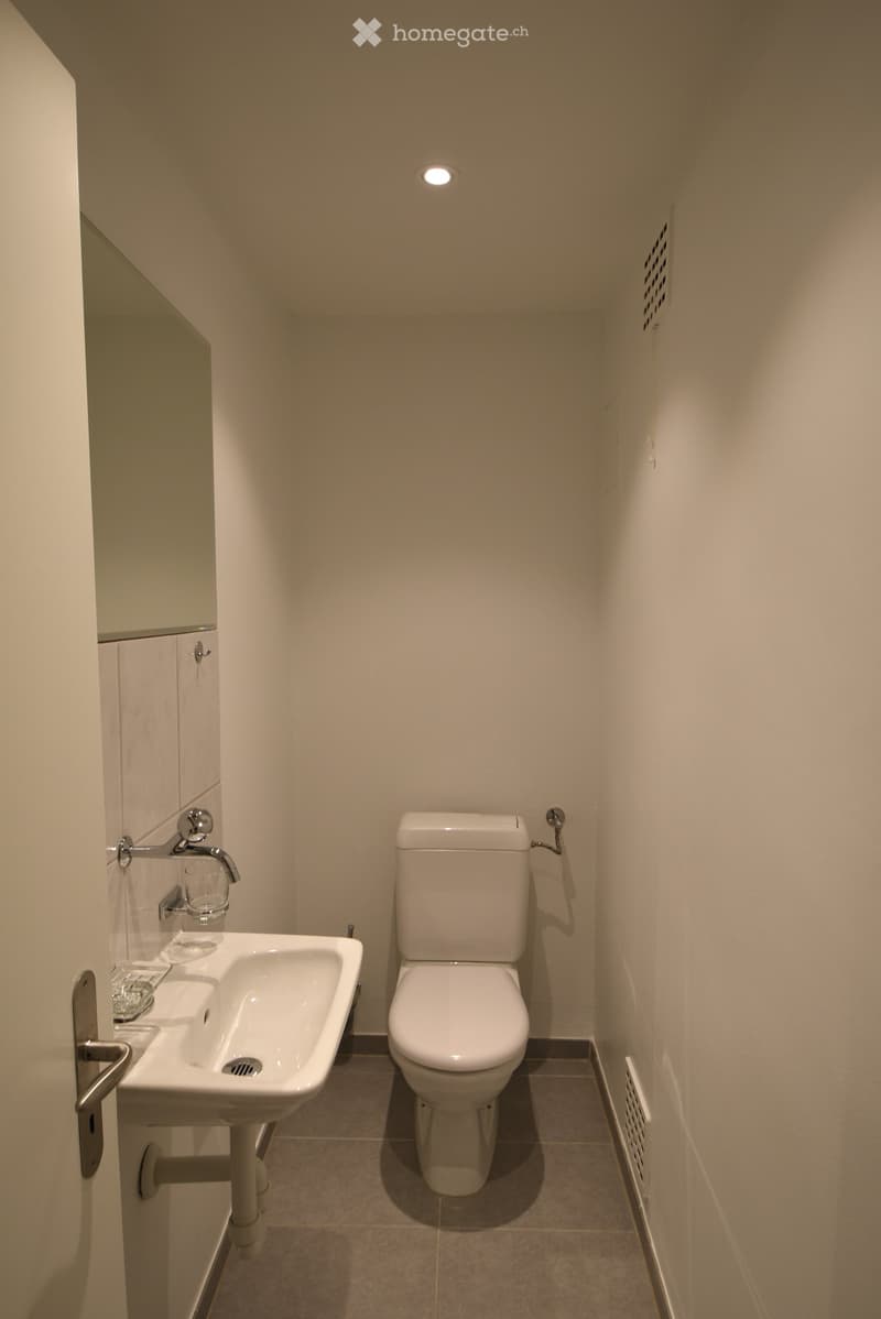 Modern bathroom with a toilet, sink, and faucet. The sink has a cup placed on it. The walls are light-colored, and the floor is tiled.