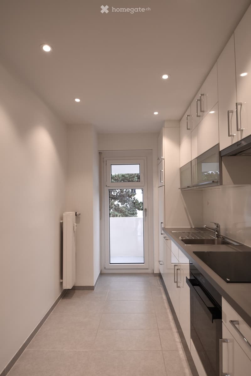 Modern kitchen with white cabinets, stainless steel sink, and built-in oven. Features recessed lighting and a large window with a view of greenery.