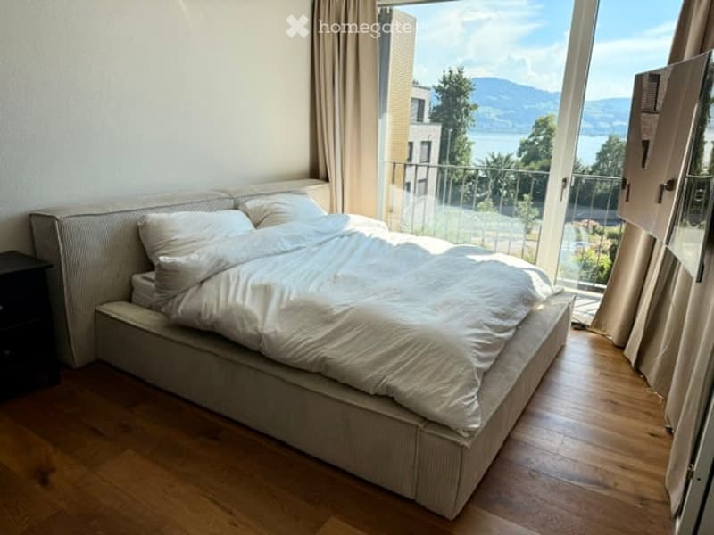 Bright bedroom with a large bed, white bedding, and a nightstand, featuring large windows with a scenic mountain view.