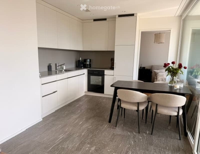 Modern kitchen with white cabinets, grey countertops, and a dining area featuring a dark wooden table and light-colored chairs. A vase with red flowers is placed on the table.