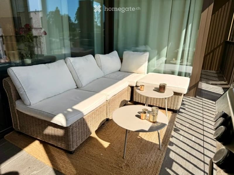 A modern outdoor seating area with a white sectional sofa, two round tables, and several small decorative items. The space features large glass windows and a wooden deck.