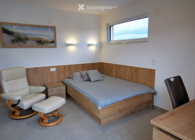 Cozy bedroom with a double bed, nightstand, white leather chair, and a small table. The room features a large window with a scenic view, a framed beach painting, and modern lighting.