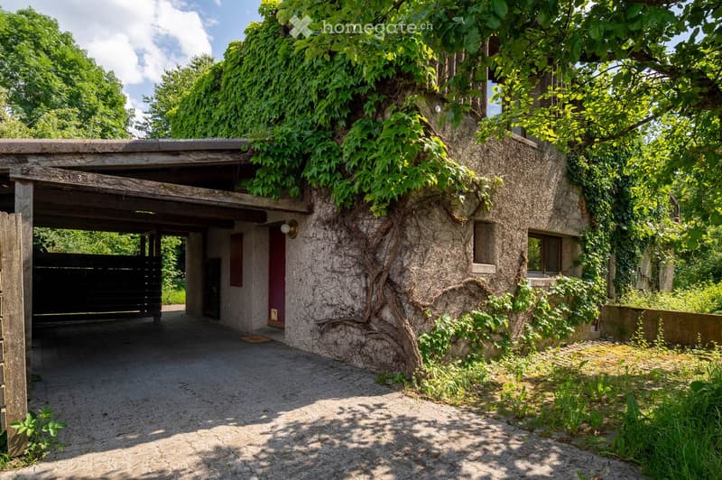 A rustic stone building with a wooden garage entrance, surrounded by lush greenery and trees. The building features a textured stone facade with climbing vines and a small porch area.