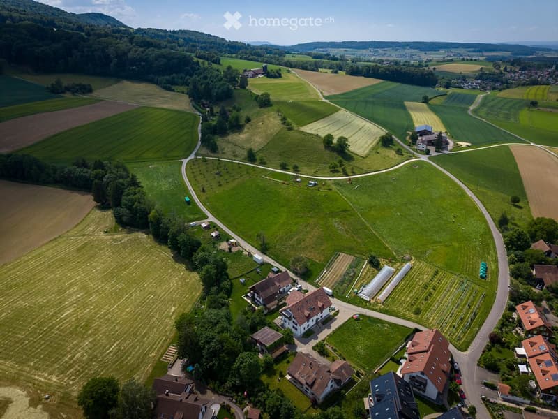 Aerial view of a rural area with several houses, green fields, and a winding road. The landscape includes cultivated farmland, trees, and a small village with scattered buildings.