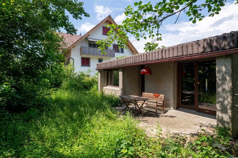 A small outdoor seating area with a wooden table and bench, attached to a modern house with a balcony and lush greenery.