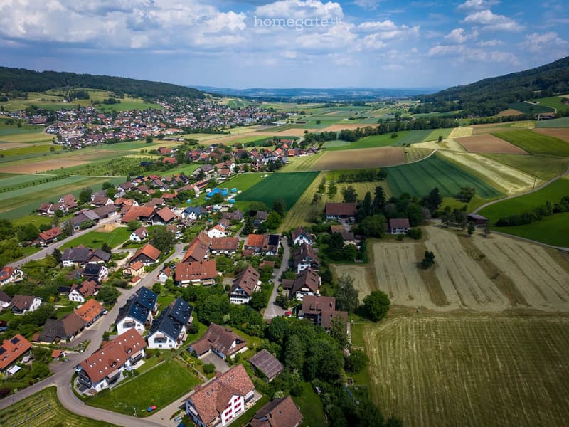 Aerial view of a small village with numerous houses, surrounded by green fields and hills under a partly cloudy sky.