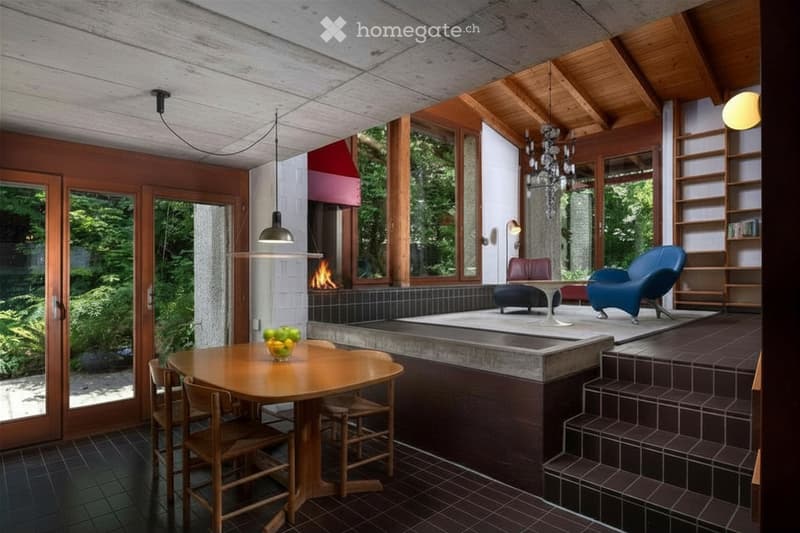 Modern living room with a fireplace, large windows, wooden table, chairs, sofa, armchair, bookshelf, and pendant lights.