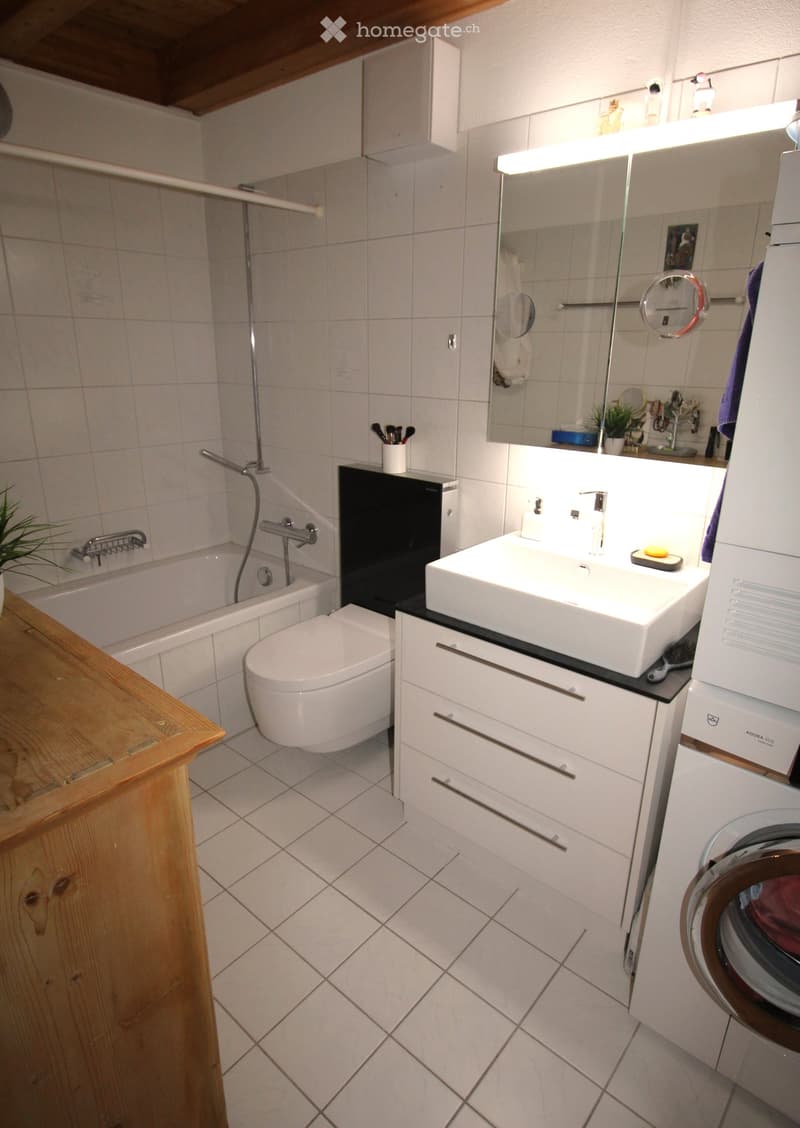 Modern bathroom with white tiles, a bathtub, toilet, sink, mirror, and a washing machine. Features include a wooden shelf and a small plant on the windowsill.