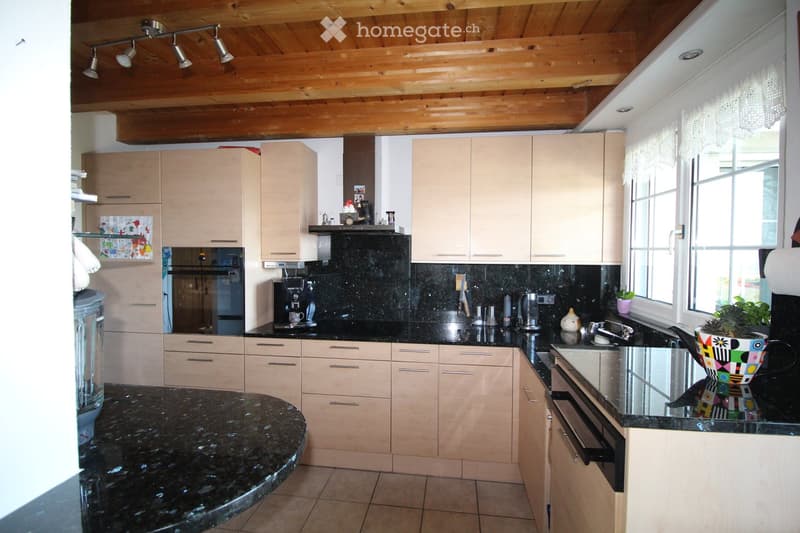 Modern kitchen with light wood cabinets, black granite countertops, stainless steel appliances, and a large island. Features include a coffee maker, blender, and decorative bowl on the counter.