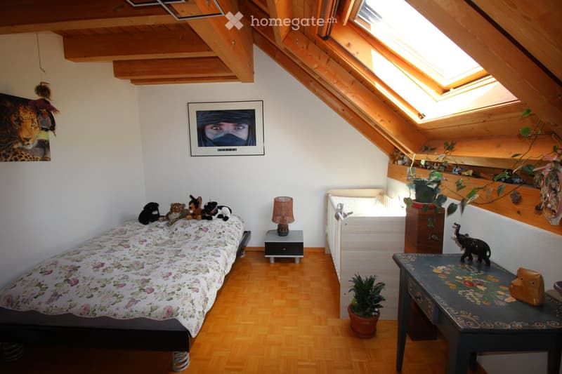 Cozy attic bedroom with wooden slanted ceilings, skylights, a single bed with floral bedding, nightstand with lamp, small table with decorative items, and potted plants.