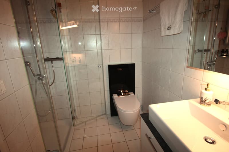 Modern bathroom with white tiles, a toilet, a sink with a mirror, a glass-enclosed shower, and a towel hanging on a rack.
