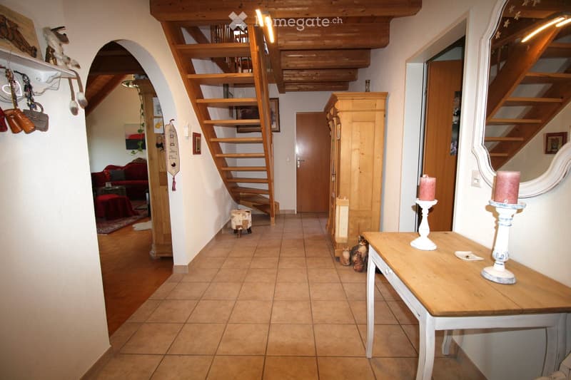 Cozy hallway with wooden staircase, tiled floor, and decorative items including a small table with candles, a cat, and a red sofa visible in the background.