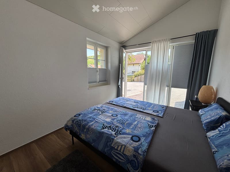 Cozy bedroom with a double bed, nightstand, and lamp. The room features wooden flooring, large windows, and a sliding door leading to an outdoor area.