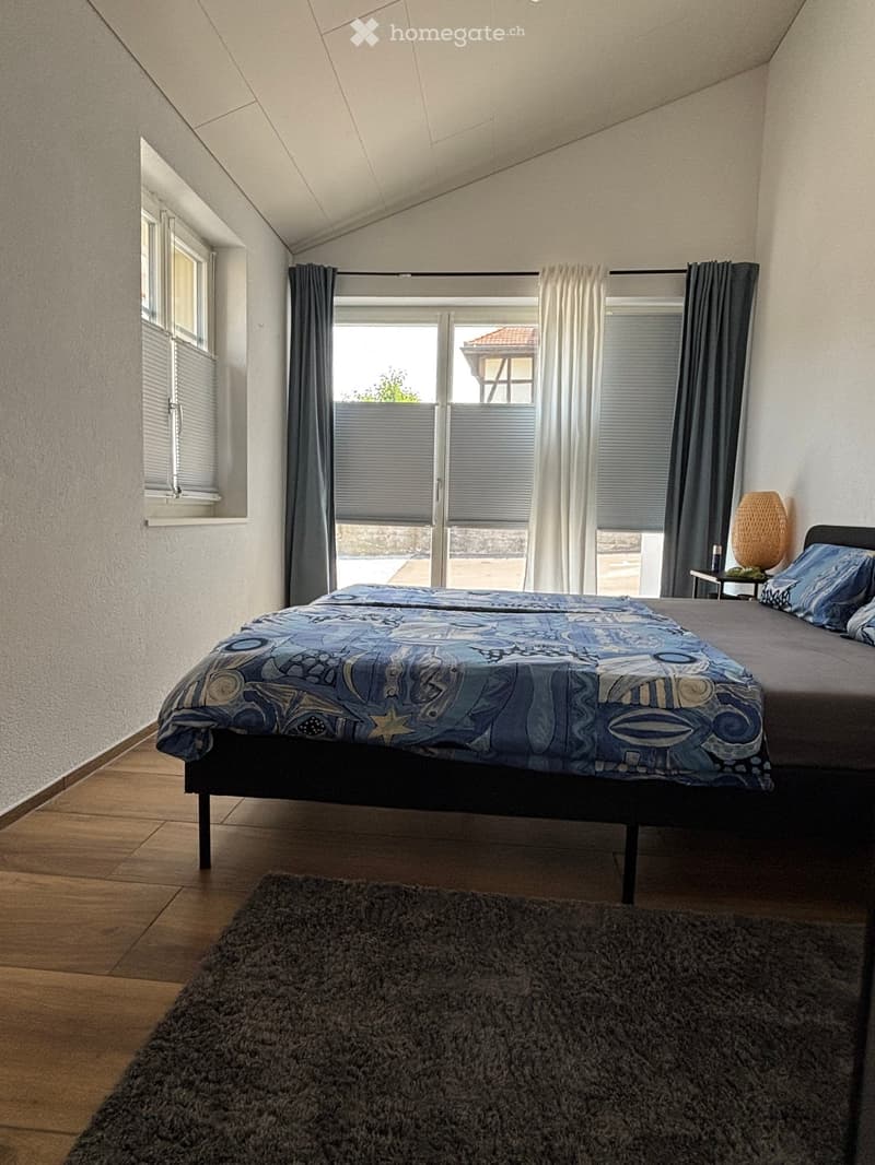 Cozy bedroom with a double bed, nightstand, lamp, and a large rug on the floor. The room features large windows with blinds and curtains, and a slanted ceiling typical of an attic space.