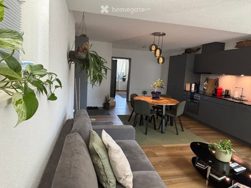 Modern open-plan living and dining area with grey sofa, coffee table, dining table with chairs, and kitchen area with cabinets and appliances.