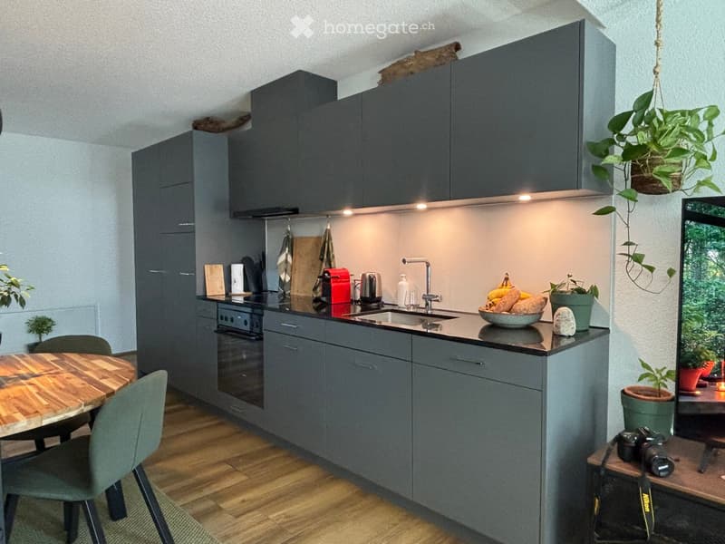 Modern kitchen with grey cabinets, wooden floor, and a small dining area. Includes a sink, oven, and various kitchen appliances.