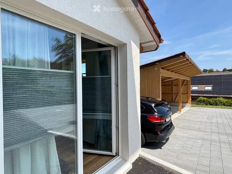 Modern house exterior with large glass sliding doors, covered wooden carport, and parked car.