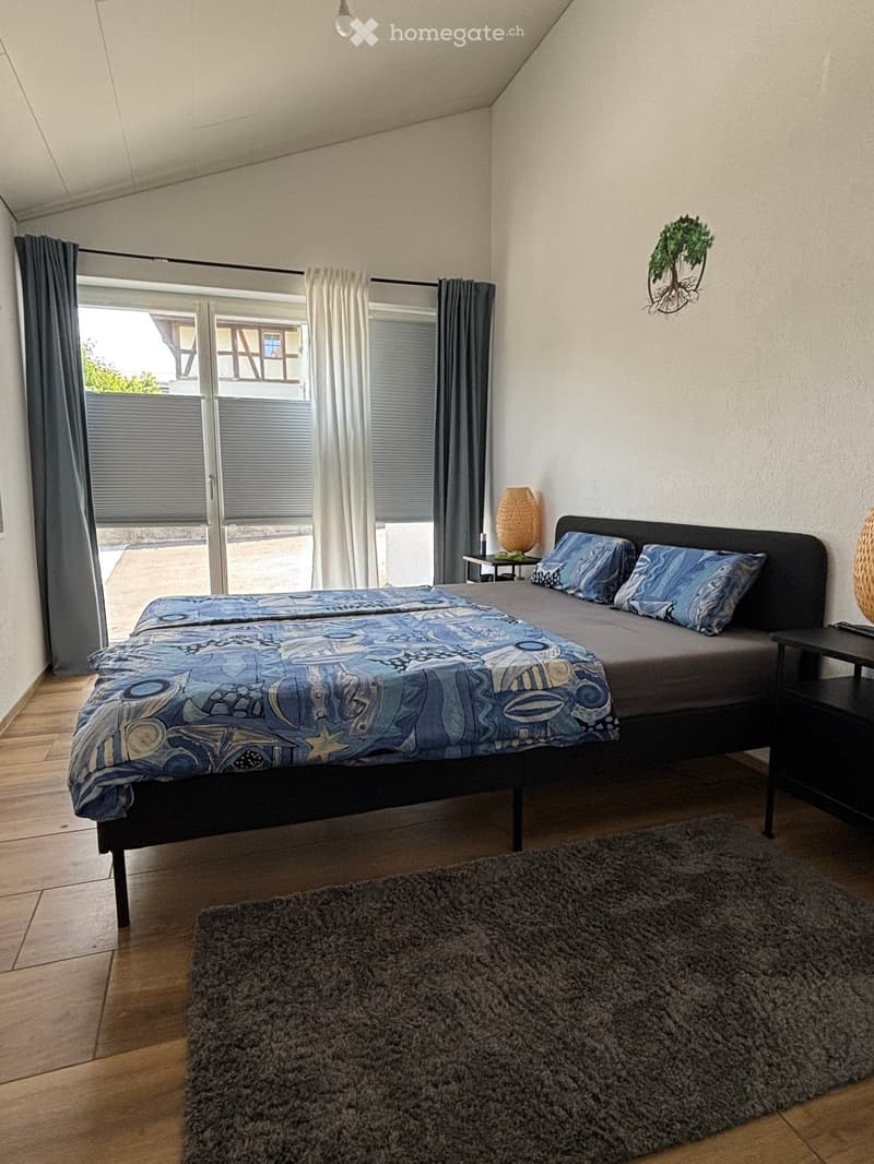 Cozy bedroom with a double bed, nightstands, a lamp, a shaggy rug, and large windows with curtains.